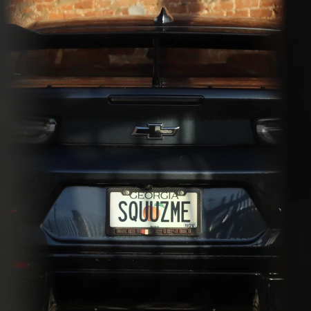 Rear view of a dark grey Chevrolet Camaro with a vanity license plate that reads 'SQUUZME', set against a textured brick wall.