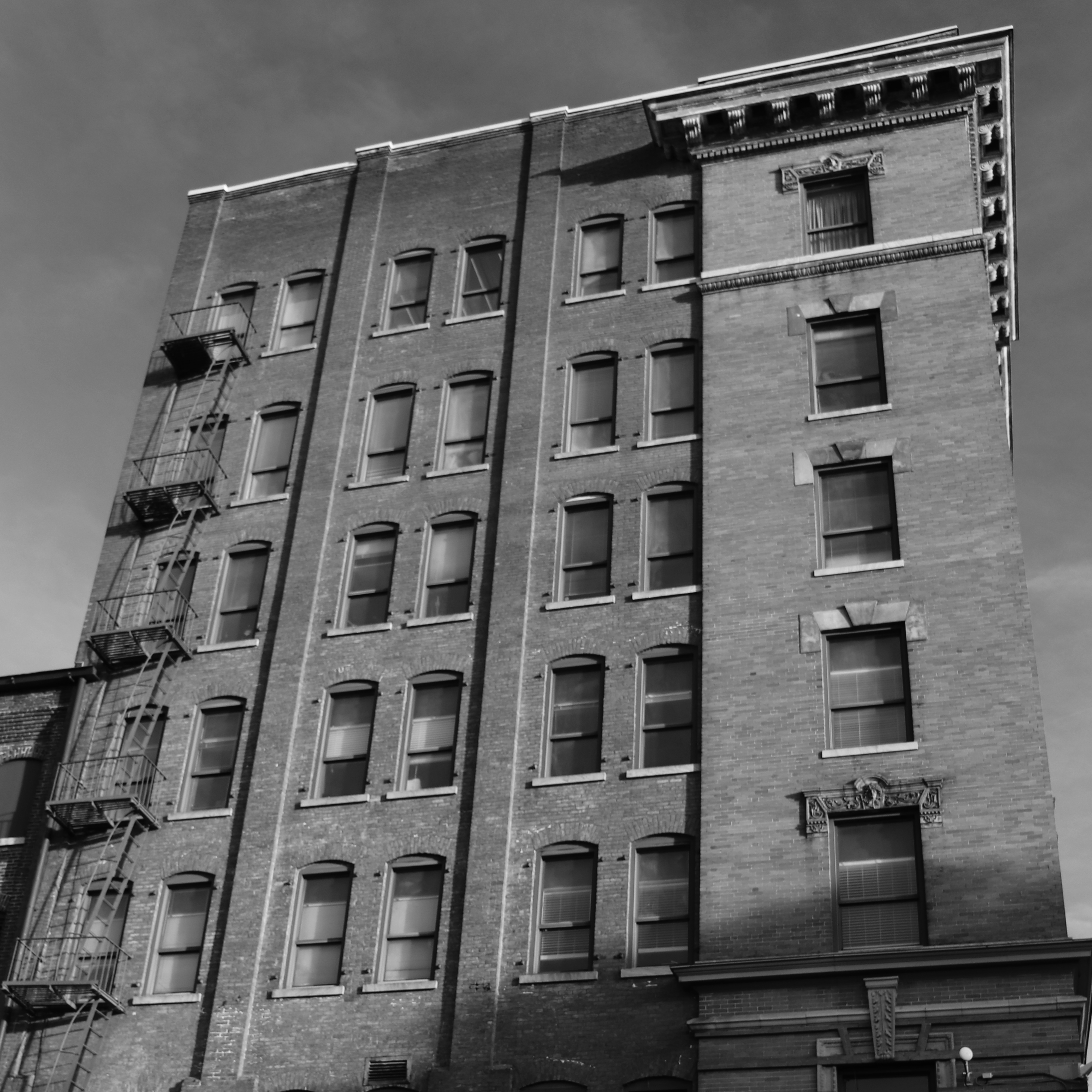 Black and white photograph of a brick apartment building exterior with numerous windows, a fire escape, and decorative architectural details.