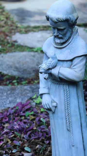 Statue of Saint Francis with a bird perched on his hand, displayed outside in a garden-like setting with stone steps.