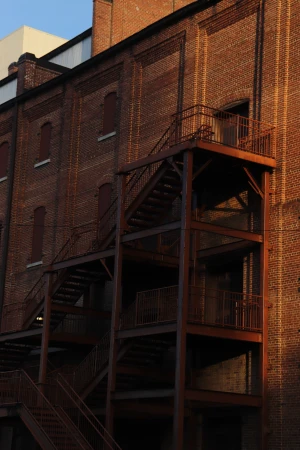 Exterior of a brick building with metal fire escape stairs under a blue sky on a sunny day, creating shadows and highlighting textures.