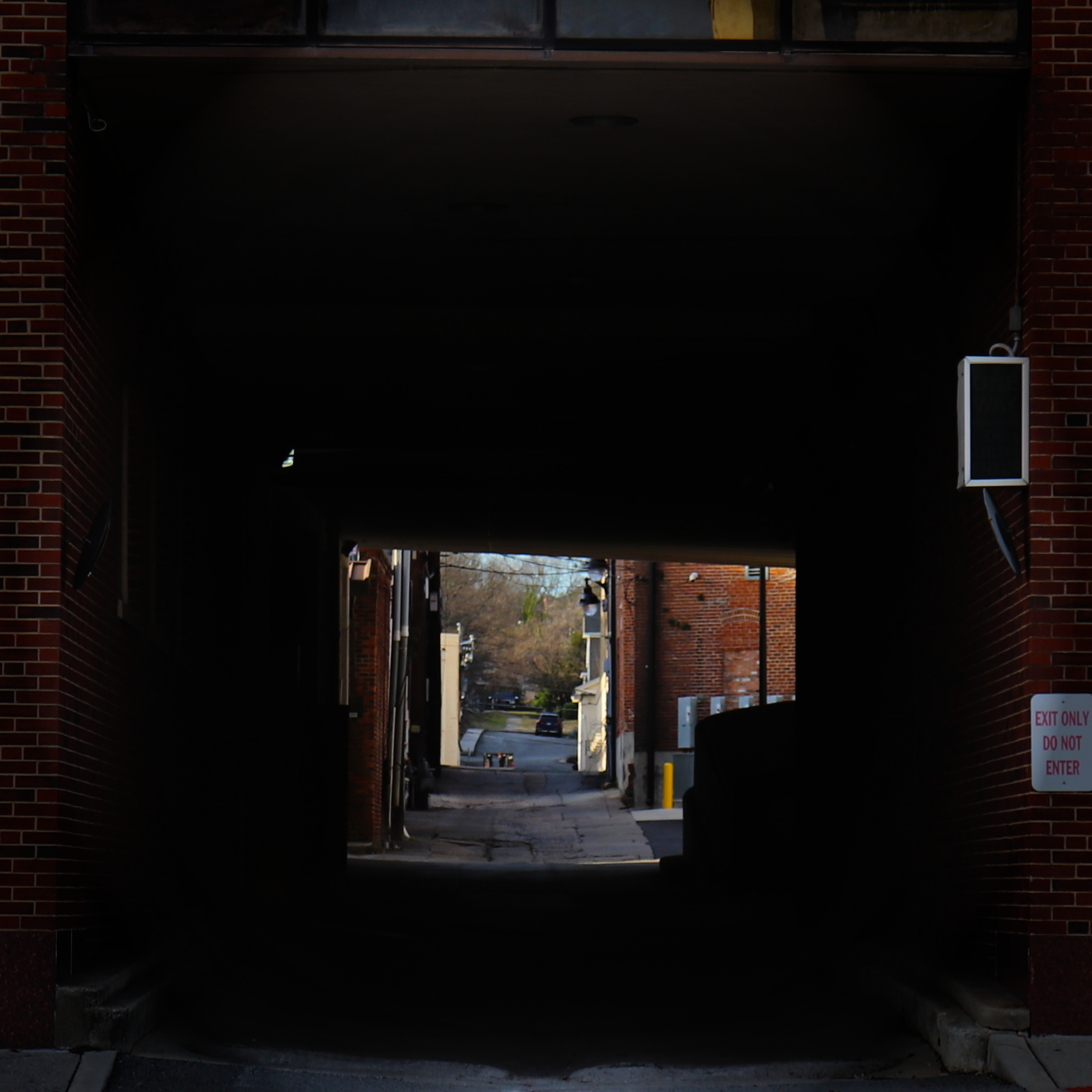 View of alleyway and road through dark brick tunnel with signage on wall. Urban streetscape with limited daylight beyond dark tunnel.