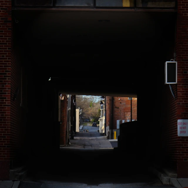 View of alleyway and road through dark brick tunnel with signage on wall. Urban streetscape with limited daylight beyond dark tunnel.