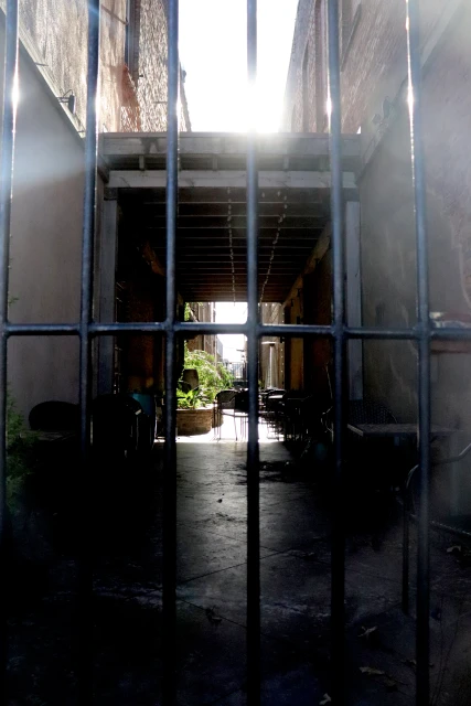 Outdoor corridor with iron bars, tables, and chairs visible through the bars, sunlight is streaming in at the end of the corridor.