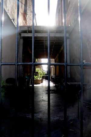 Outdoor corridor with iron bars, tables, and chairs visible through the bars, sunlight is streaming in at the end of the corridor.