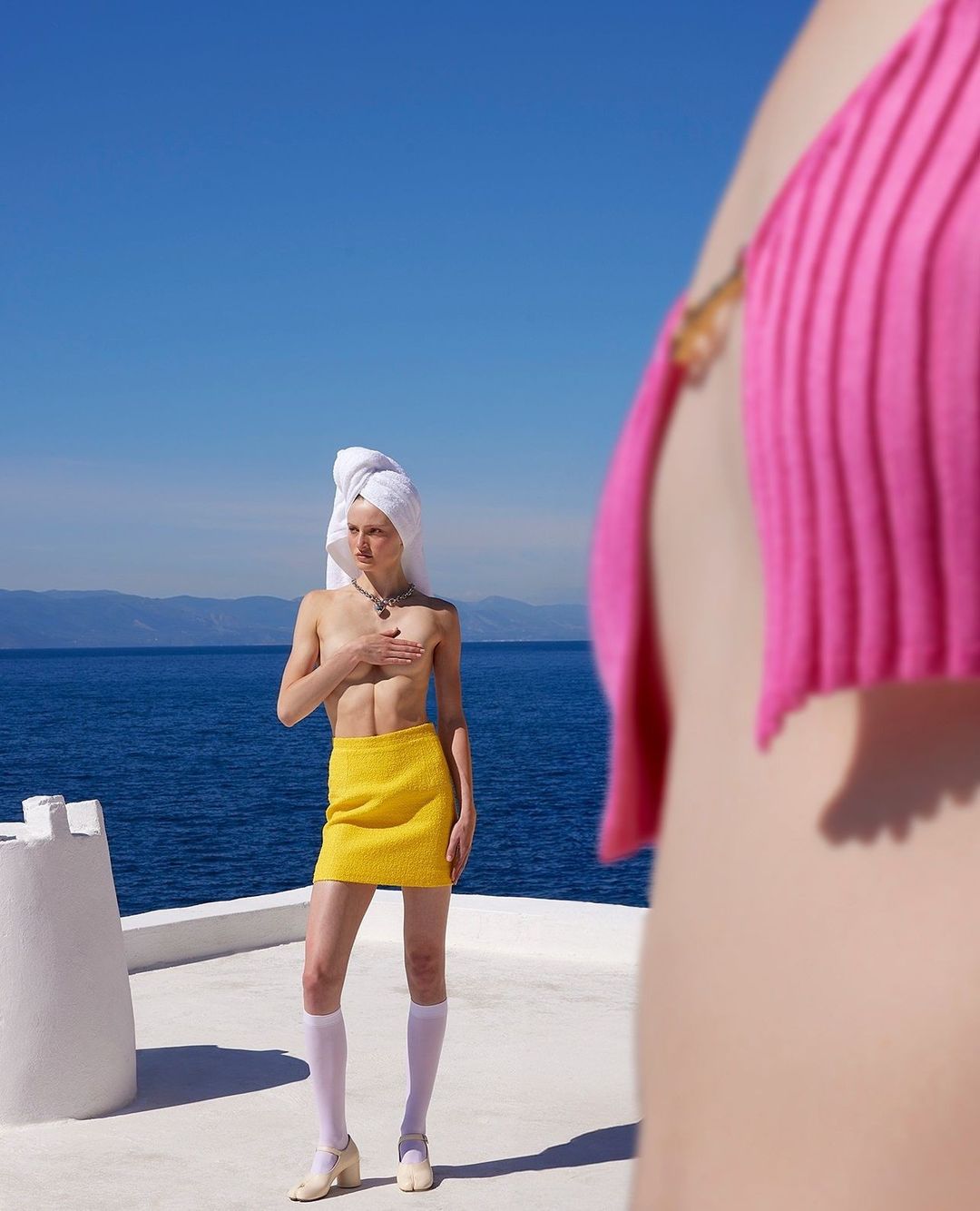 Young woman with a towel on her head, wearing a yellow skirt and knee-high socks, poses outdoors on a bright day near the ocean.