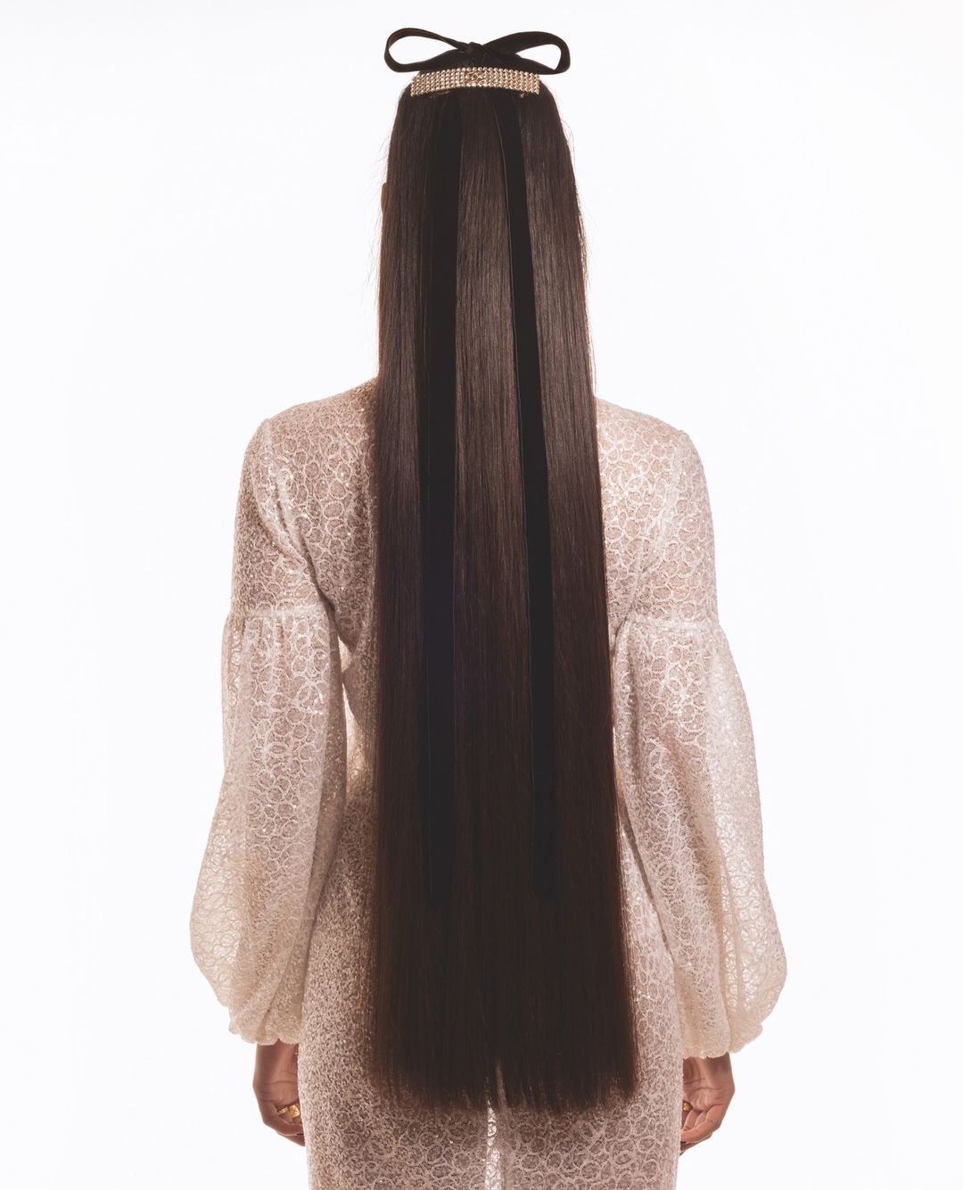 Back view of a woman with long dark brown hair styled with a bejeweled headband and a floral-patterned top against a white backdrop.