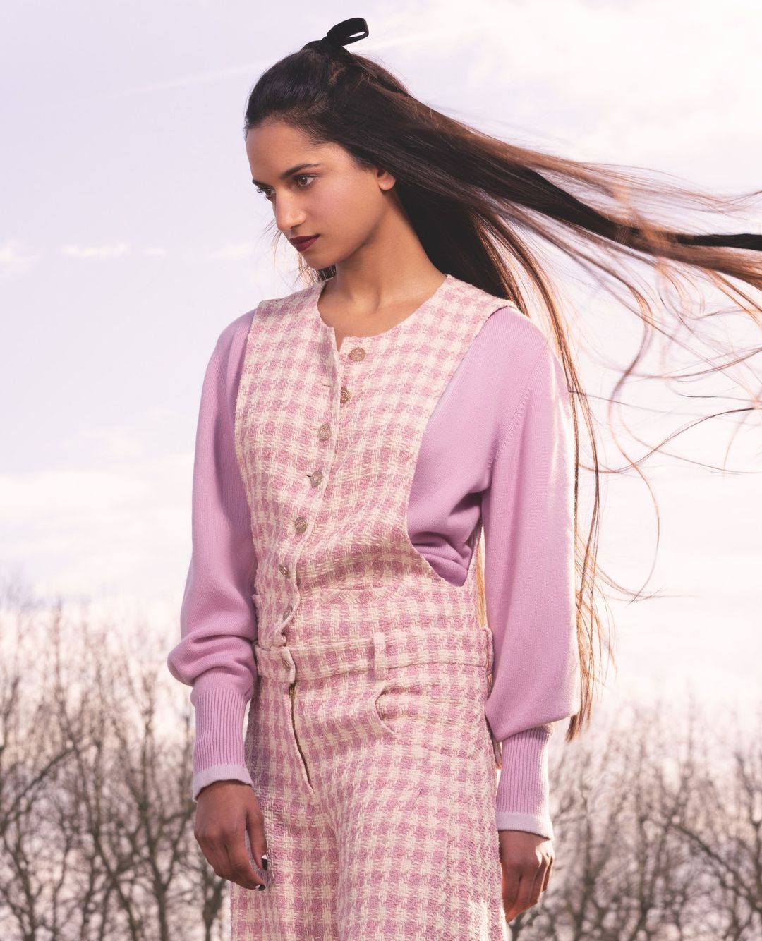 Young woman in a pink patterned jumpsuit and sweater poses outdoors against a soft sky. Her long hair blows in the breeze.