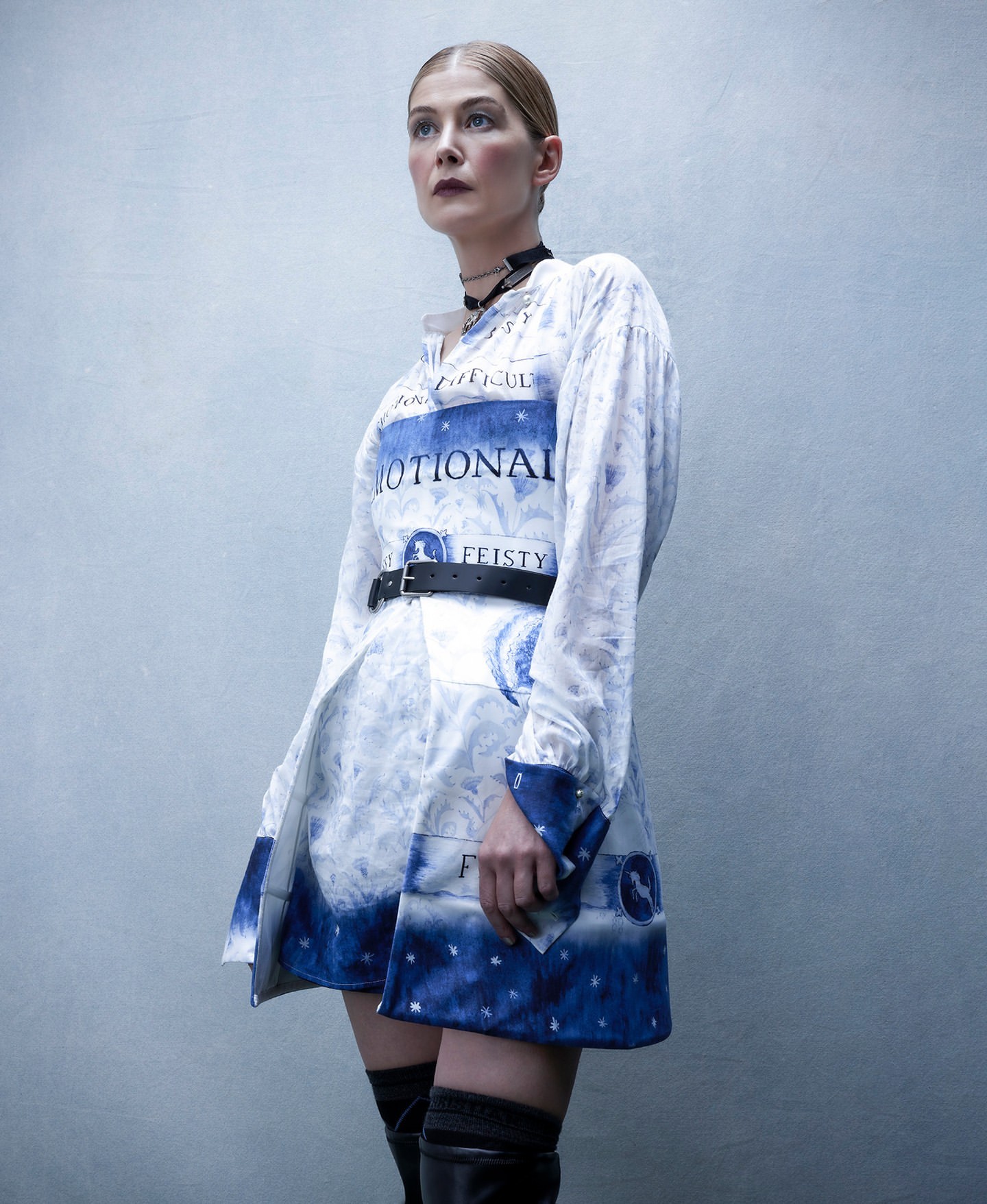 Woman in literary-themed dress with black belt and thigh-high boots poses indoors with a smooth gradient studio background.