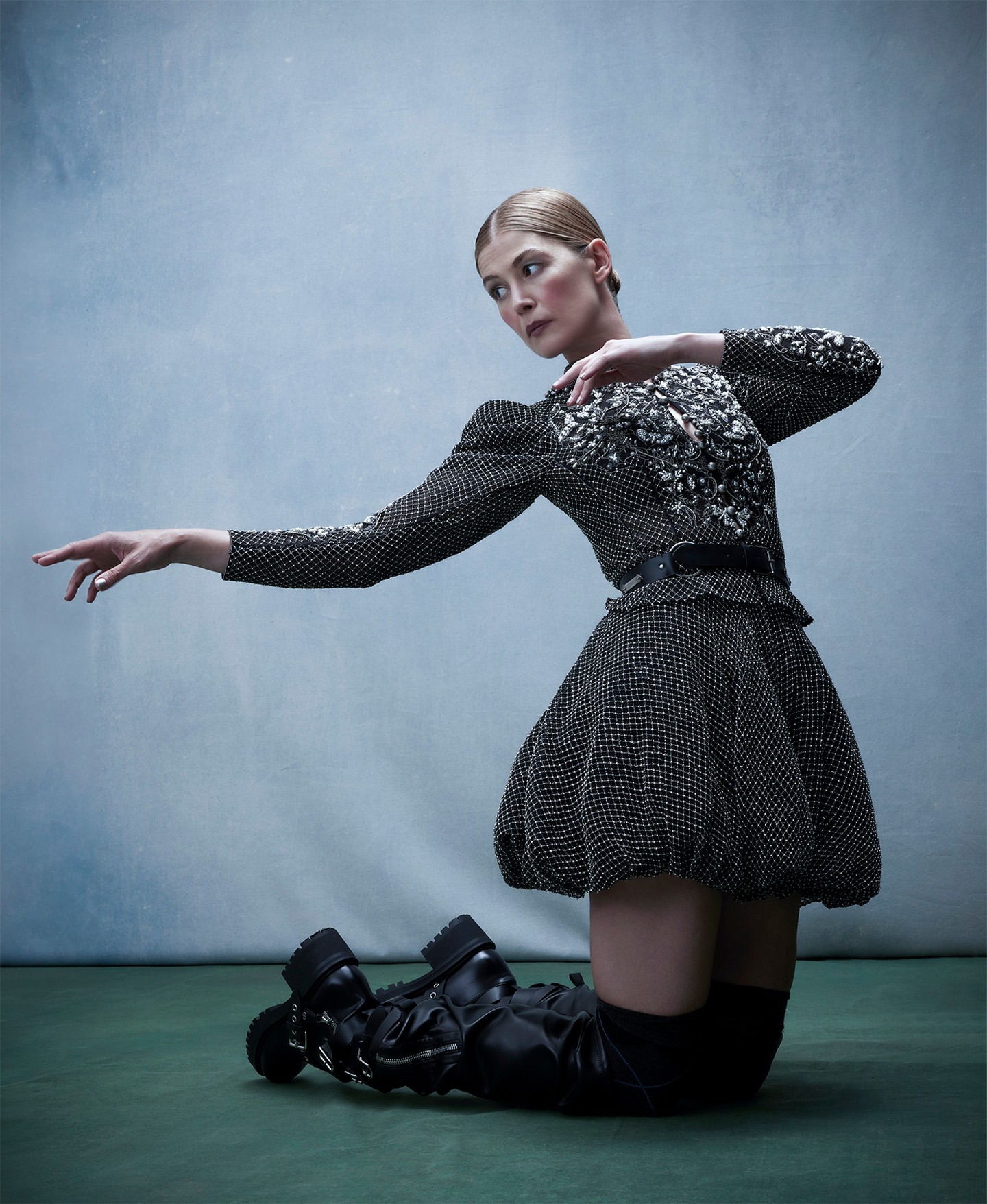 A woman poses in a short black and white spotted dress with sleeves and black boots on a green surface in front of a mottled blue backdrop.