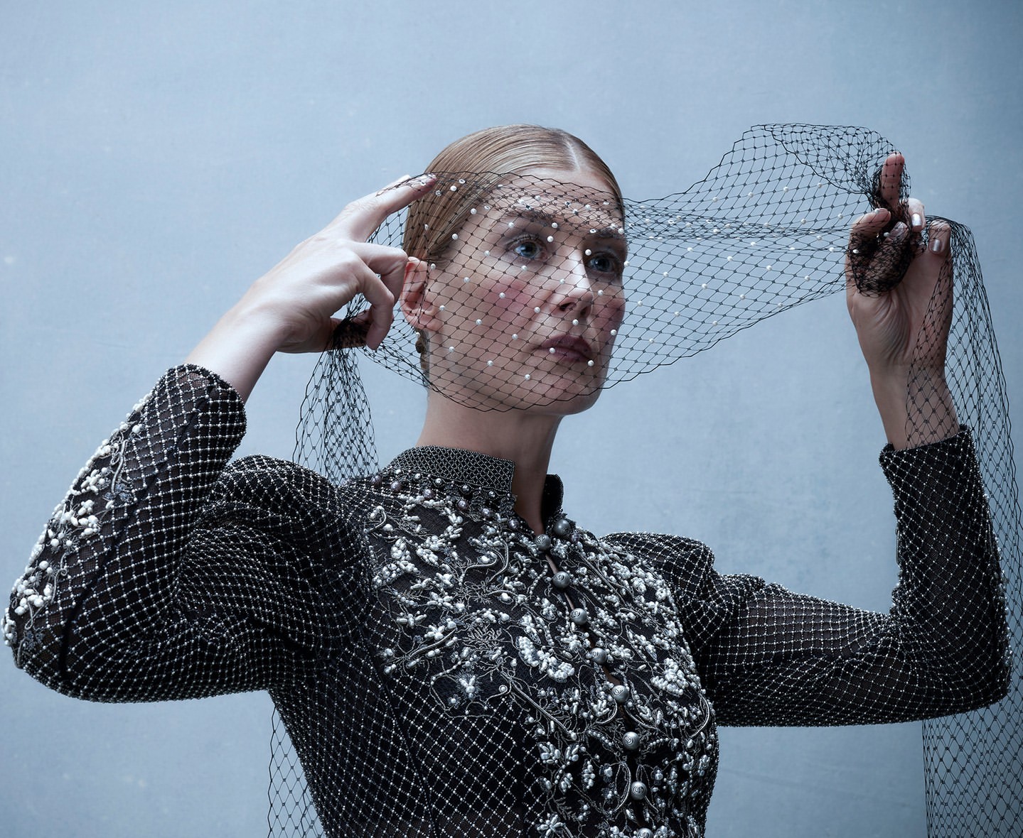 Woman in an embellished high-neck top and beaded netting over her head, posing against a neutral gradient background, studio shot.