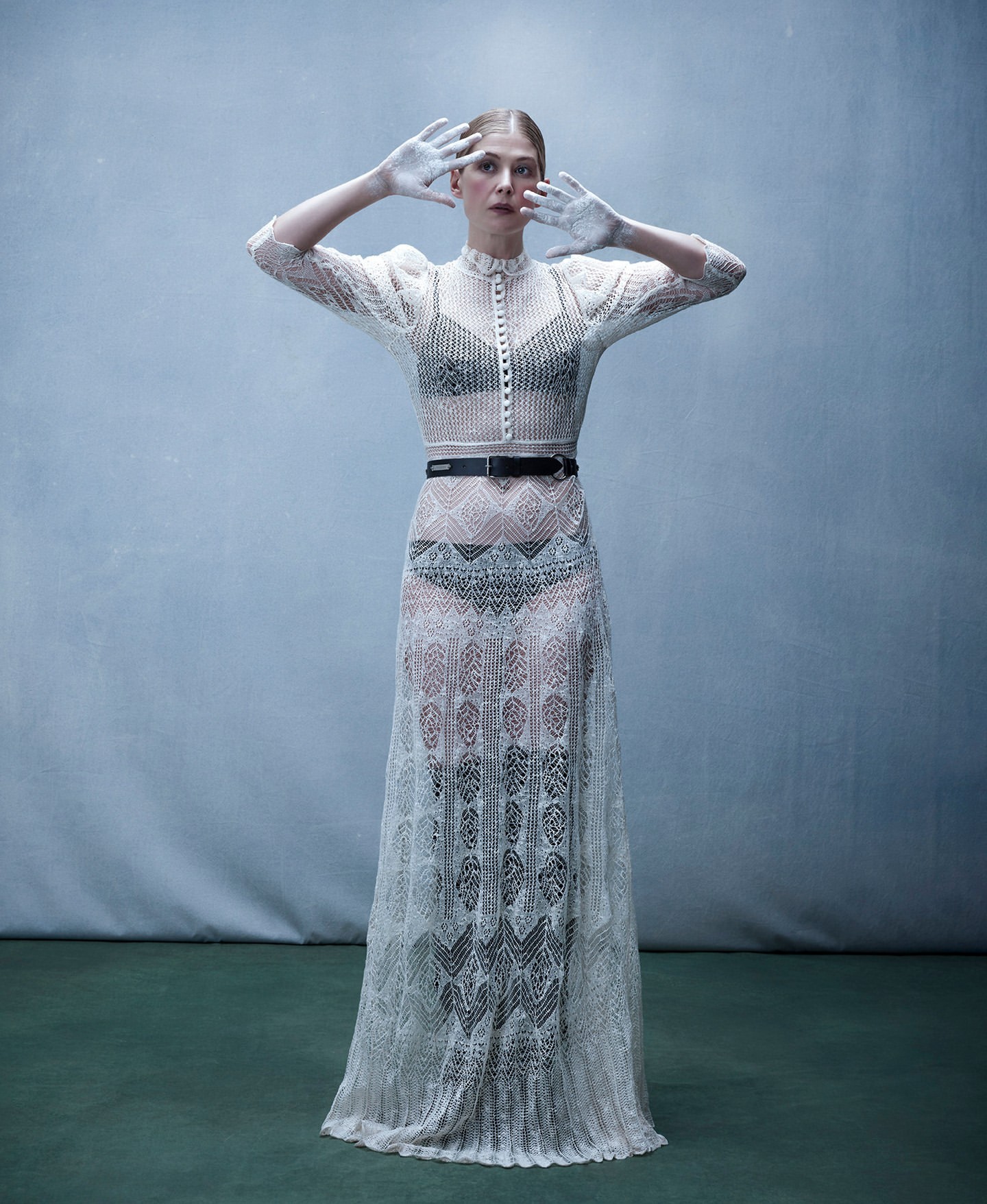 Woman in a sheer white lace dress stands with painted hands up in a studio, creating a high-fashion editorial image. 