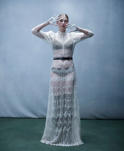 Woman in a sheer white lace dress stands with painted hands up in a studio, creating a high-fashion editorial image. 