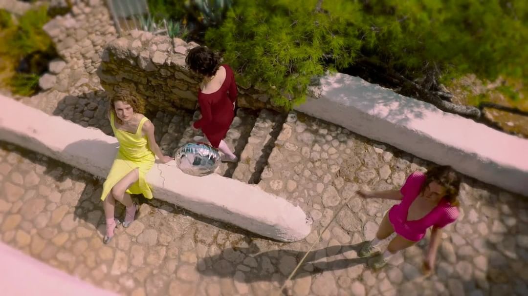 Three women in dresses pose on a stone patio next to a disco ball, captured from a high angle in bright sunlight.
