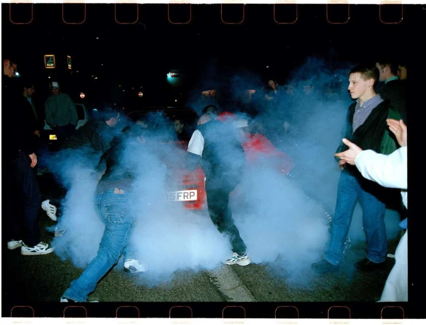People gather around a red car enveloped in thick white smoke at night, with buildings and streetlights visible in the background.