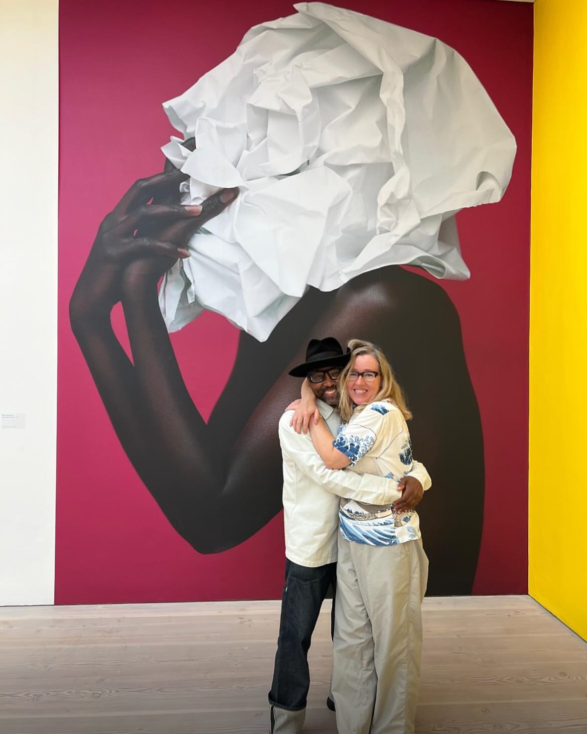 Two people embrace in front of a large art piece. One is a man wearing a hat, jeans, and a white jacket. The other is a woman with glasses.
