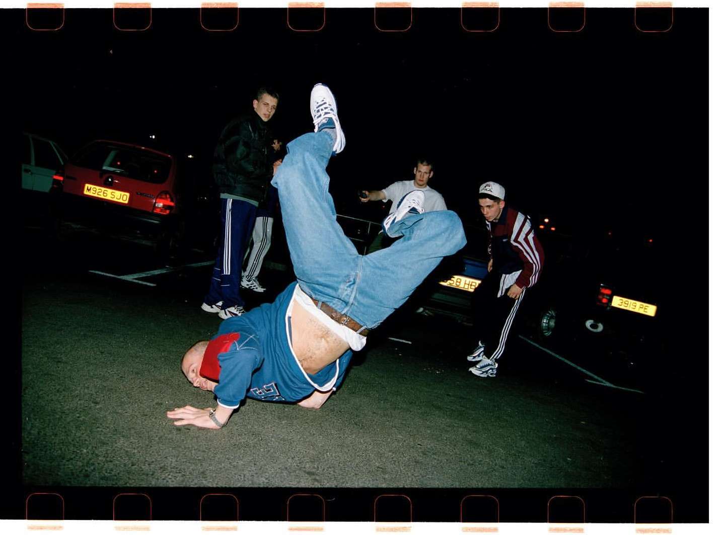 Young adult breakdancer performing a handstand outdoors at night, surrounded by a group of other young adults near parked cars.