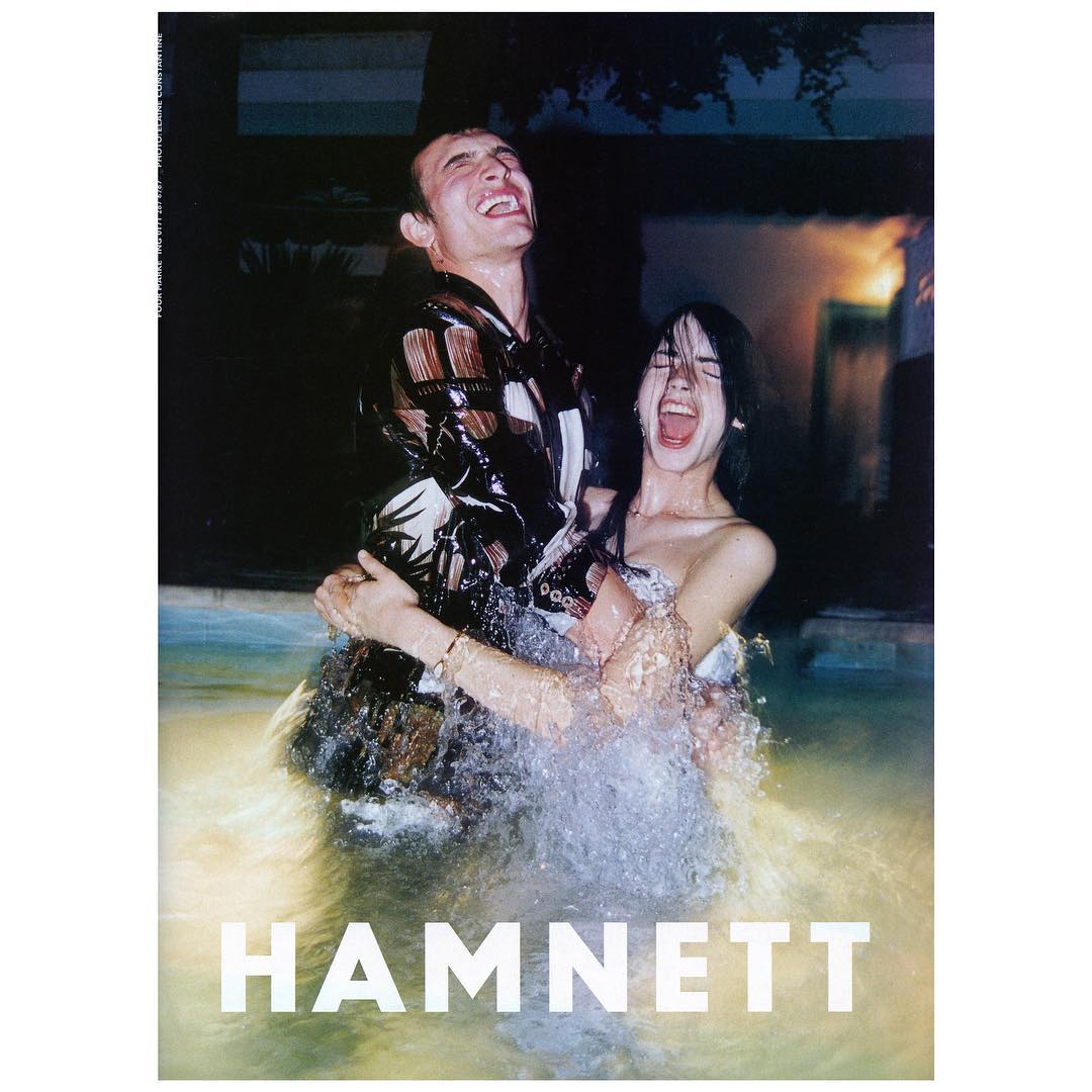 A couple in a pool smile with joy, water splashing as they embrace. The man wears a patterned shirt and the woman is in a white dress.