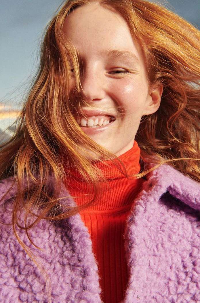 Smiling woman with red hair wears orange turtleneck and lilac coat, captured in a close-up outdoor shot with diffused natural light.