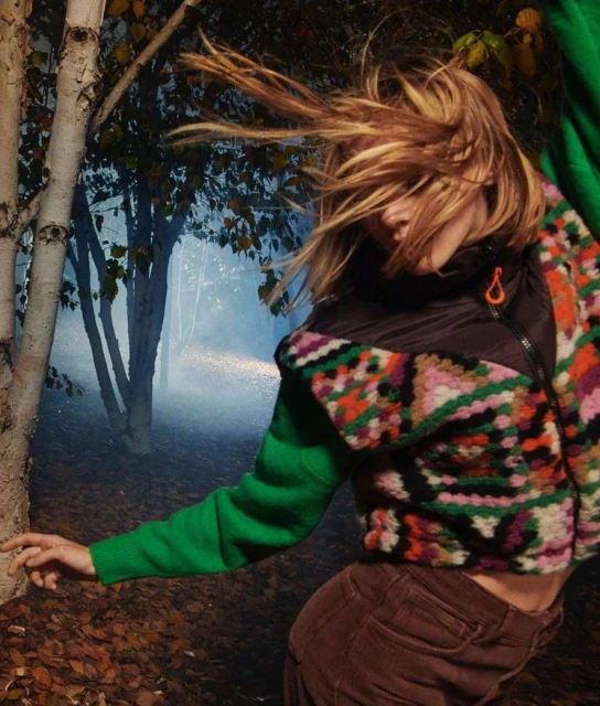 A fair-skinned woman with blonde hair poses in an outdoor setting with birch trees and a misty background, wearing a colorful patterned jacket.