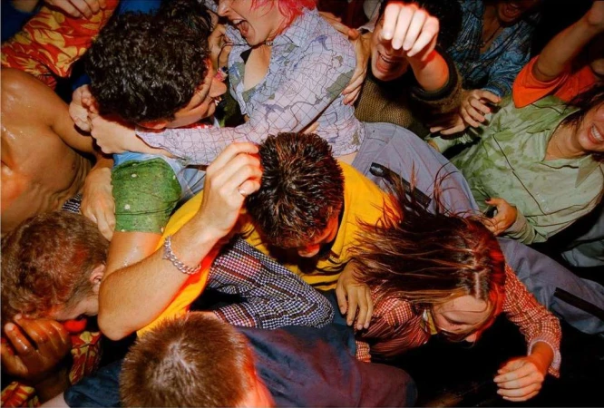 Dynamic crowd scene with multiple people, some embracing and laughing in a dimly lit indoor setting with varied expressions and clothing.