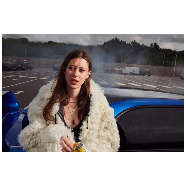 Adult woman in a furry coat stands by a blue car in a parking lot with an open bag of chips; smoke is visible in the background.