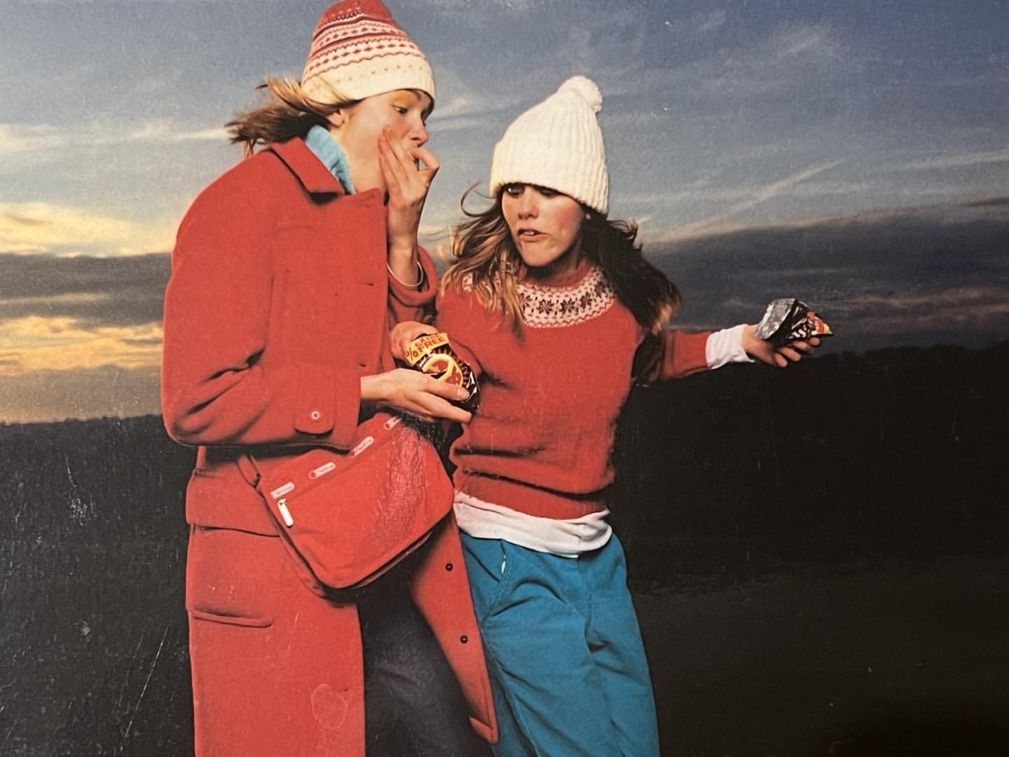 Two young women in winter hats and colorful sweaters hold snacks outdoors against a dusky sky, dressed for a casual winter day.