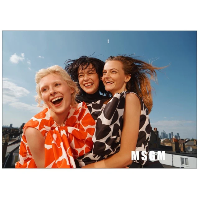 Three women smile outdoors in fashionable tops with heart patterns against a blue sky and cityscape backdrop. Fashion editorial.