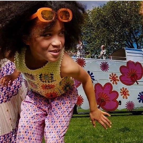 A child with afro hair and orange sunglasses poses outdoors wearing a colorful crochet top and patterned pants, with a flower-painted van behind.