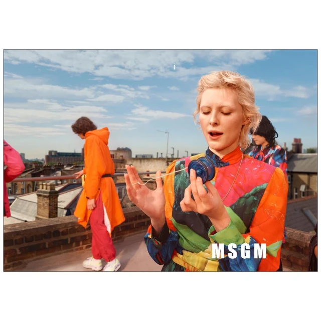 Fashion shot of a blonde adult female playing with a blue yoyo on a rooftop with other people in the background on a sunny day.