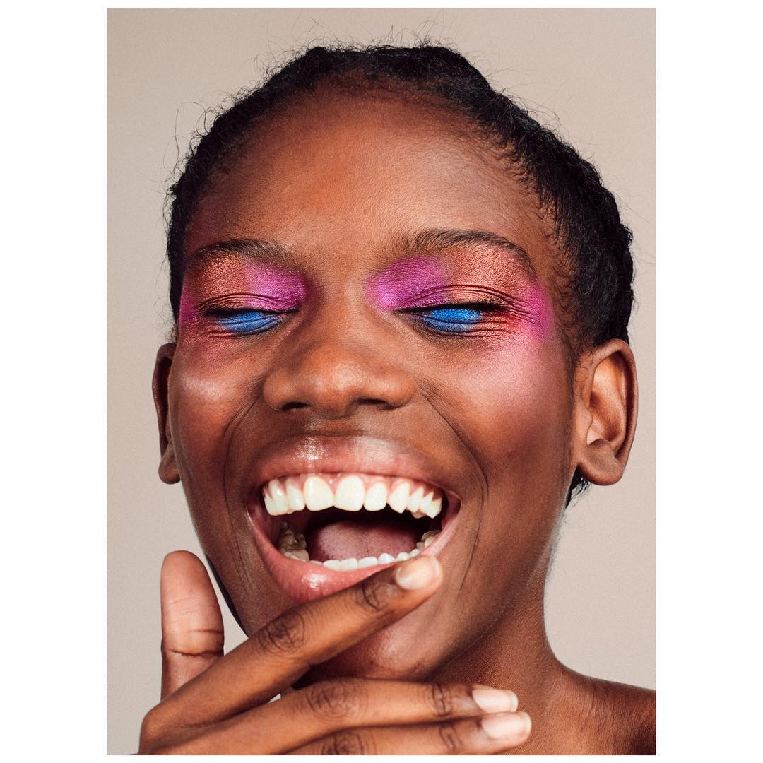 Close-up shot of an African American woman with bright pink and blue eyeshadow smiling with a finger to her face in a beauty context.