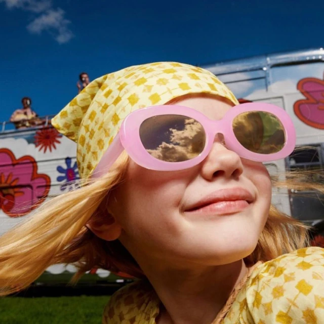 Close-up of a child wearing pink sunglasses and a patterned yellow headscarf, posing near a retro RV under a bright blue sky.