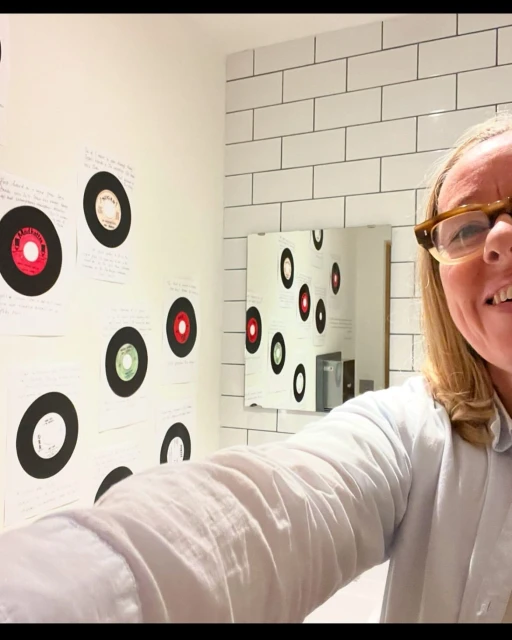 Blonde woman in glasses smiles in selfie with bathroom decorated with records on walls and mirror for a fun, musical aesthetic.