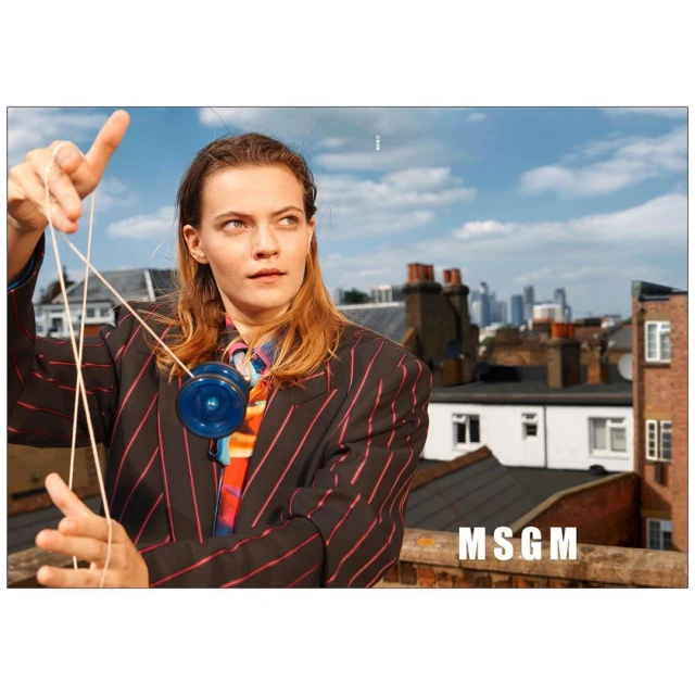 Young woman in striped blazer poses with a blue yoyo on a rooftop with a city skyline backdrop under a partly cloudy sky in daytime.