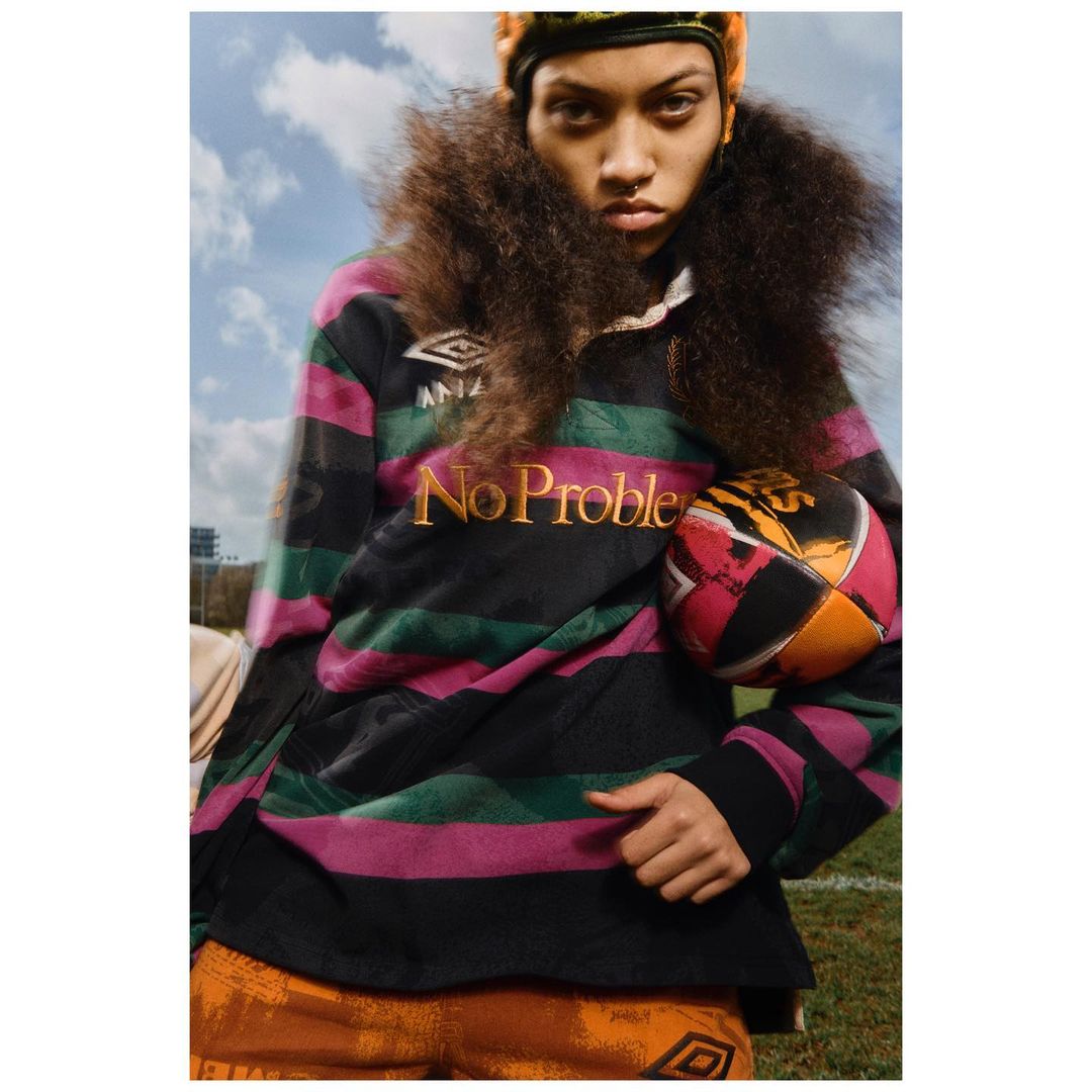 Woman in striped rugby shirt poses outdoors with ball. Headband, afro hairstyle, blue sky and field add to sporty, fashion-forward image.