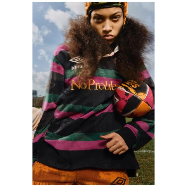 Woman in striped rugby shirt poses outdoors with ball. Headband, afro hairstyle, blue sky and field add to sporty, fashion-forward image.
