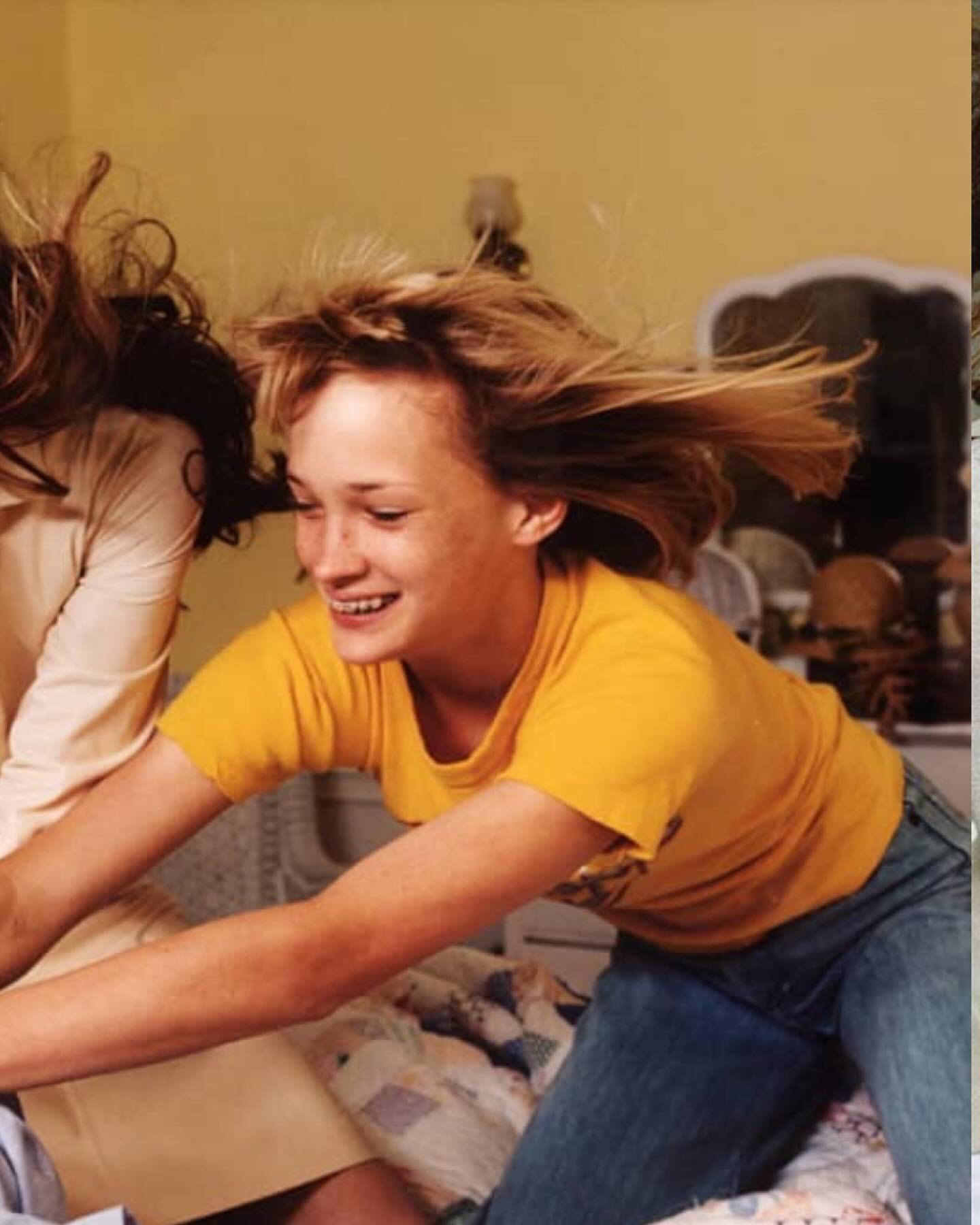 Two young women playfully jumping on a bed in an indoor bedroom with yellow walls and a white bedspread and comforter.
