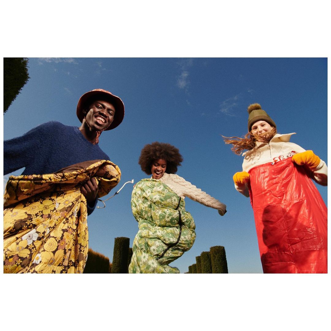 Three people in sleeping bags stand outside against a clear blue sky with green topiary in the background and a worm's eye view.