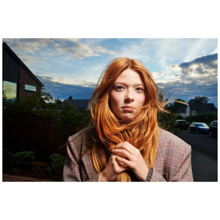 Young woman with long red hair wearing a plaid blazer outdoors under a cloudy sky in London.