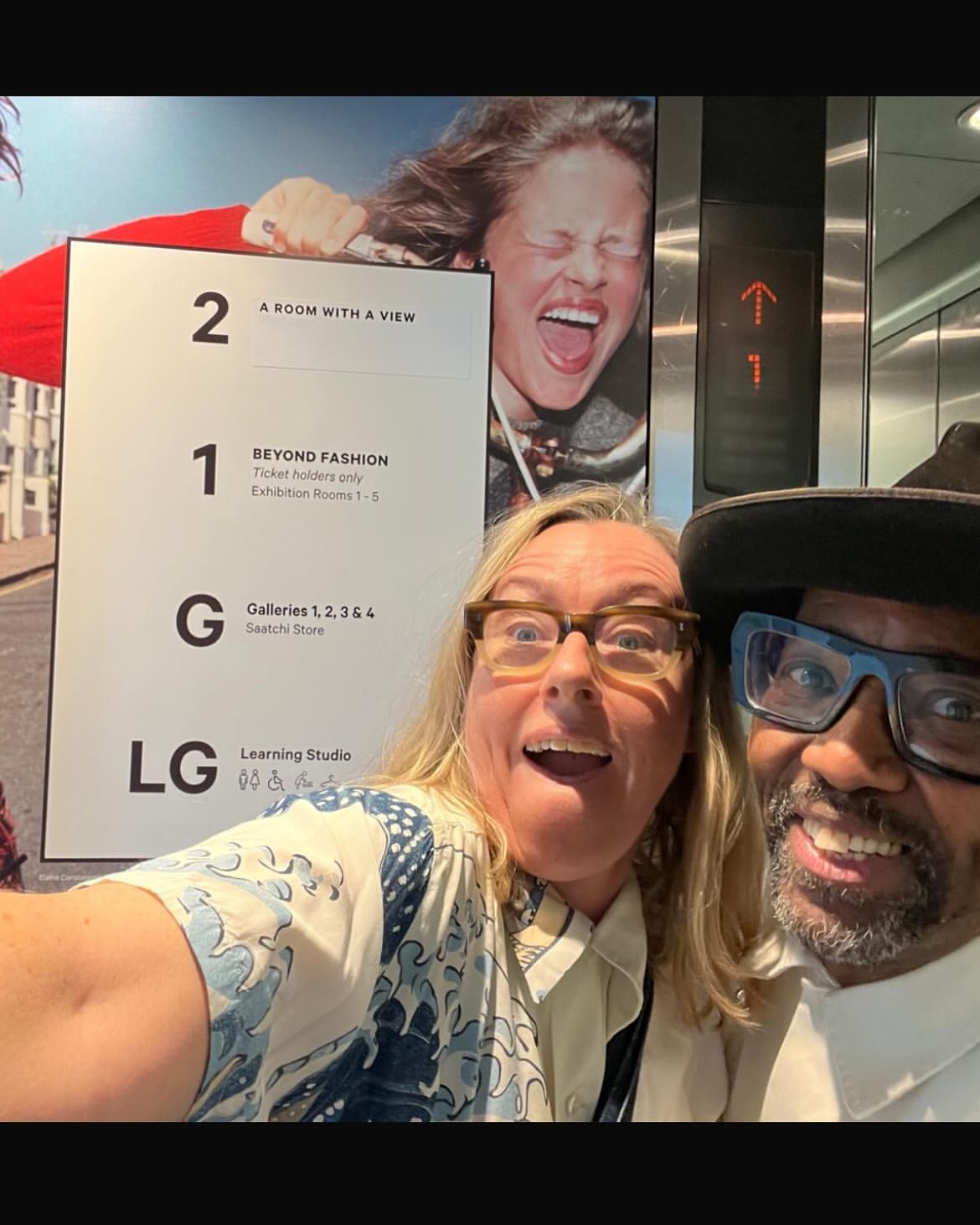 Selfie with two adults at the fashion exhibit, posing in front of an advertisement, sign for exhibition rooms, and a poster featuring model.