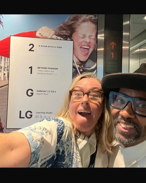 Selfie with two adults at the fashion exhibit, posing in front of an advertisement, sign for exhibition rooms, and a poster featuring model.