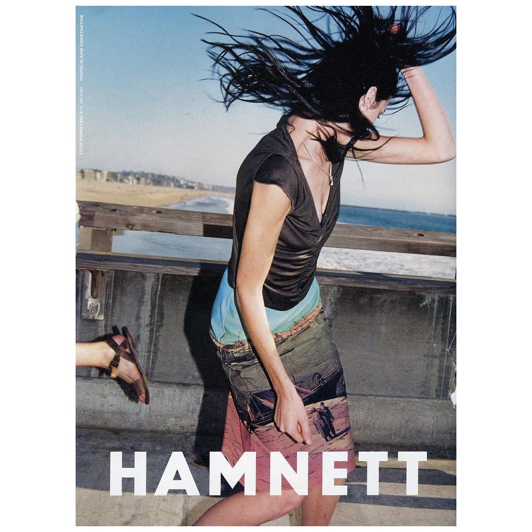 A fashion advertisement featuring a woman with wind-blown hair wearing a black top and colorful skirt on a wooden boardwalk near a beach.