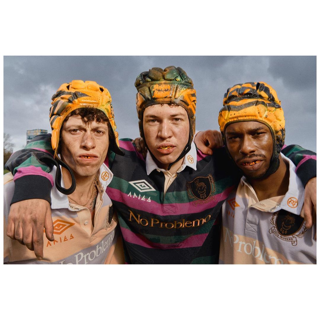 Three young adult males are seen from the chest up wearing athletic uniforms and headgear, standing outdoors under a cloudy sky.
