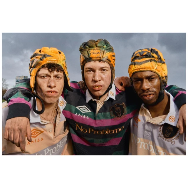 Three young adult males are seen from the chest up wearing athletic uniforms and headgear, standing outdoors under a cloudy sky.