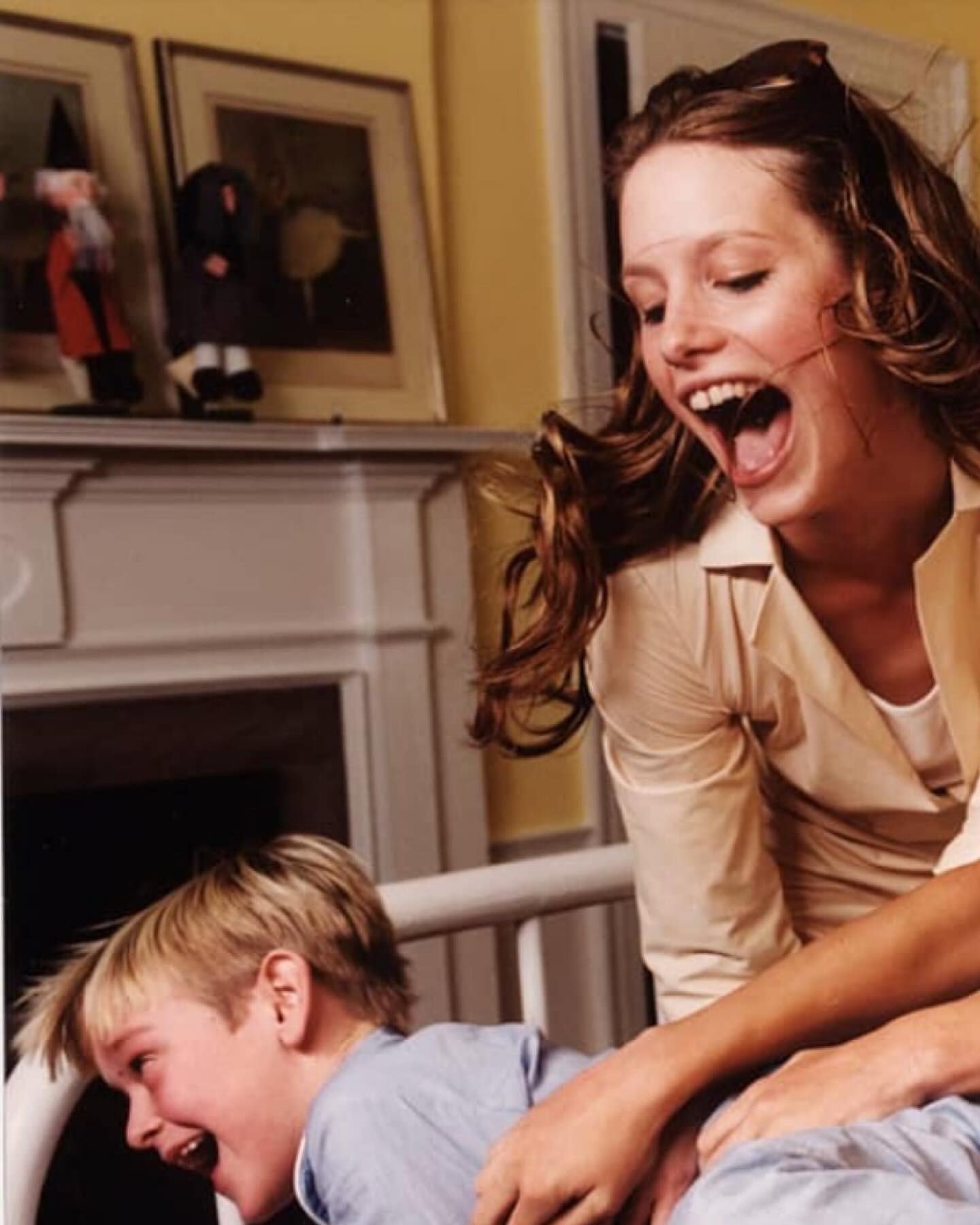 A mother and child share a laugh indoors; the woman smiles broadly with the child laughing, both appearing joyful in a bright indoor setting.