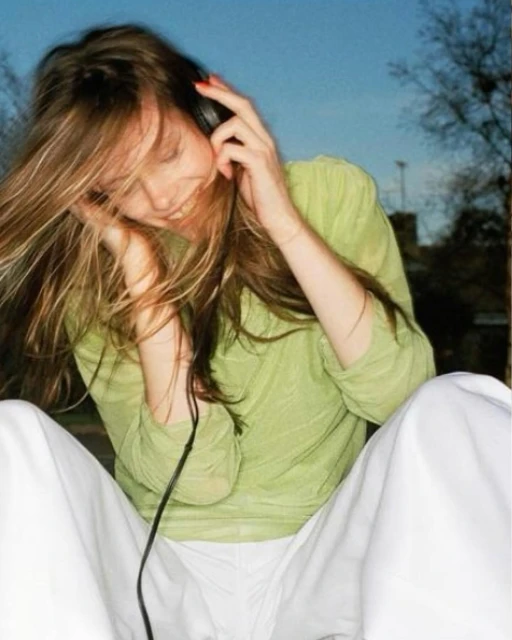 Young woman with long brown hair sits outside wearing a green top and white pants, listening to music with black headphones on her head.