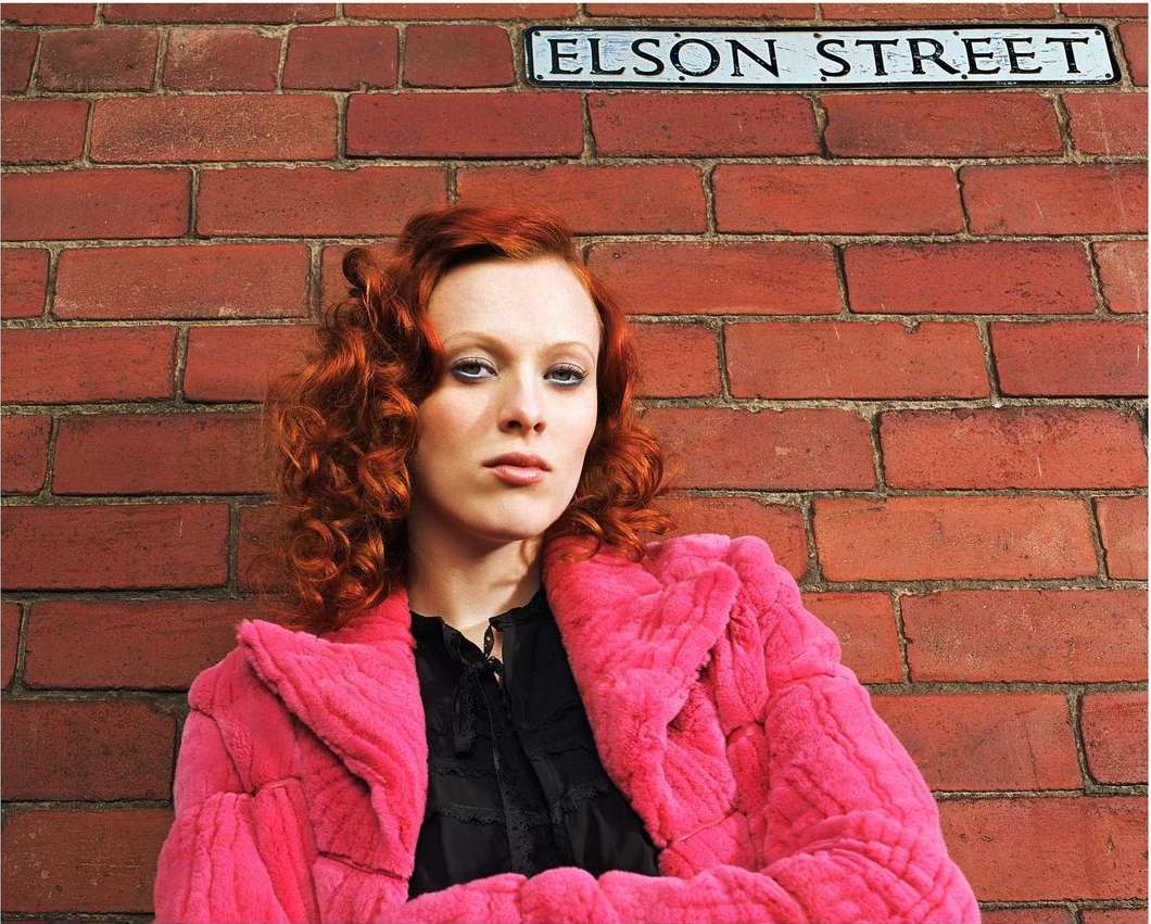 Woman with red hair wearing a pink faux fur coat standing in front of a brick wall with an Elson Street sign in the background.