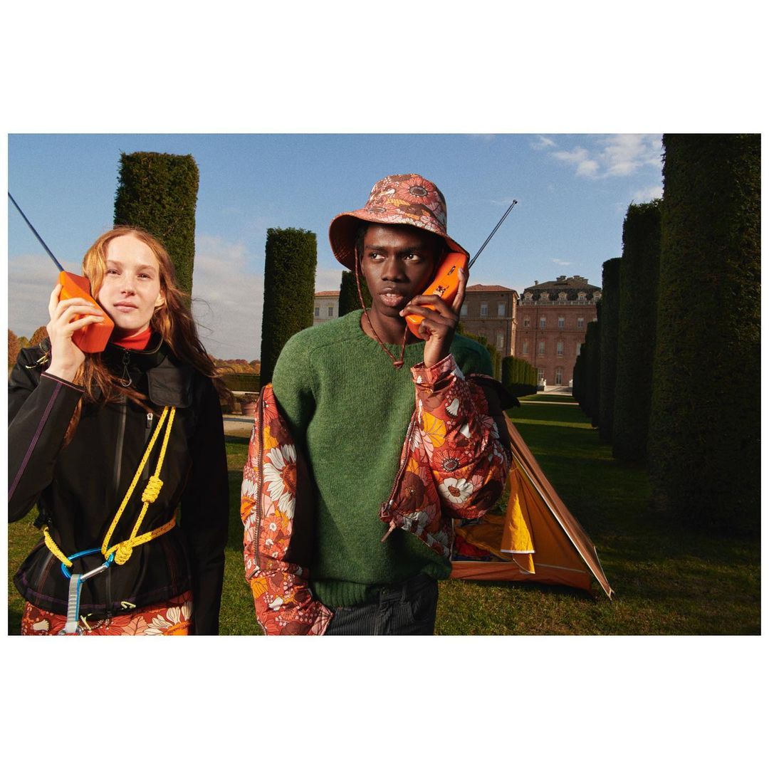 Two people in outdoor setting with a tent, wearing floral patterned outfits and holding orange brick cell phones up to their ears.