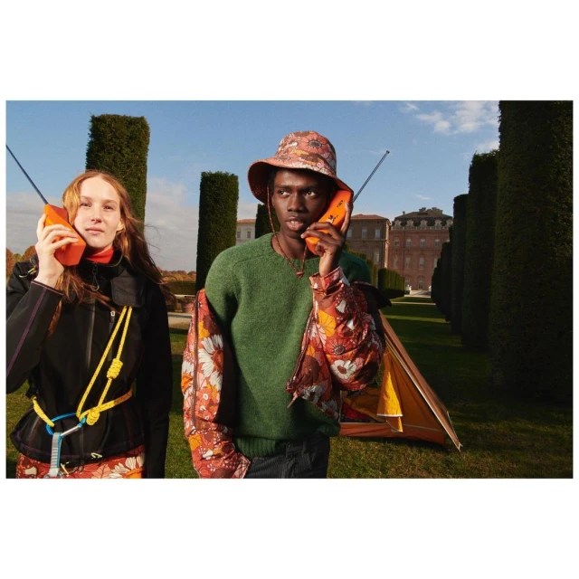 Two people in outdoor setting with a tent, wearing floral patterned outfits and holding orange brick cell phones up to their ears.