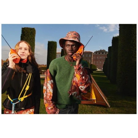 Two people in outdoor setting with a tent, wearing floral patterned outfits and holding orange brick cell phones up to their ears.