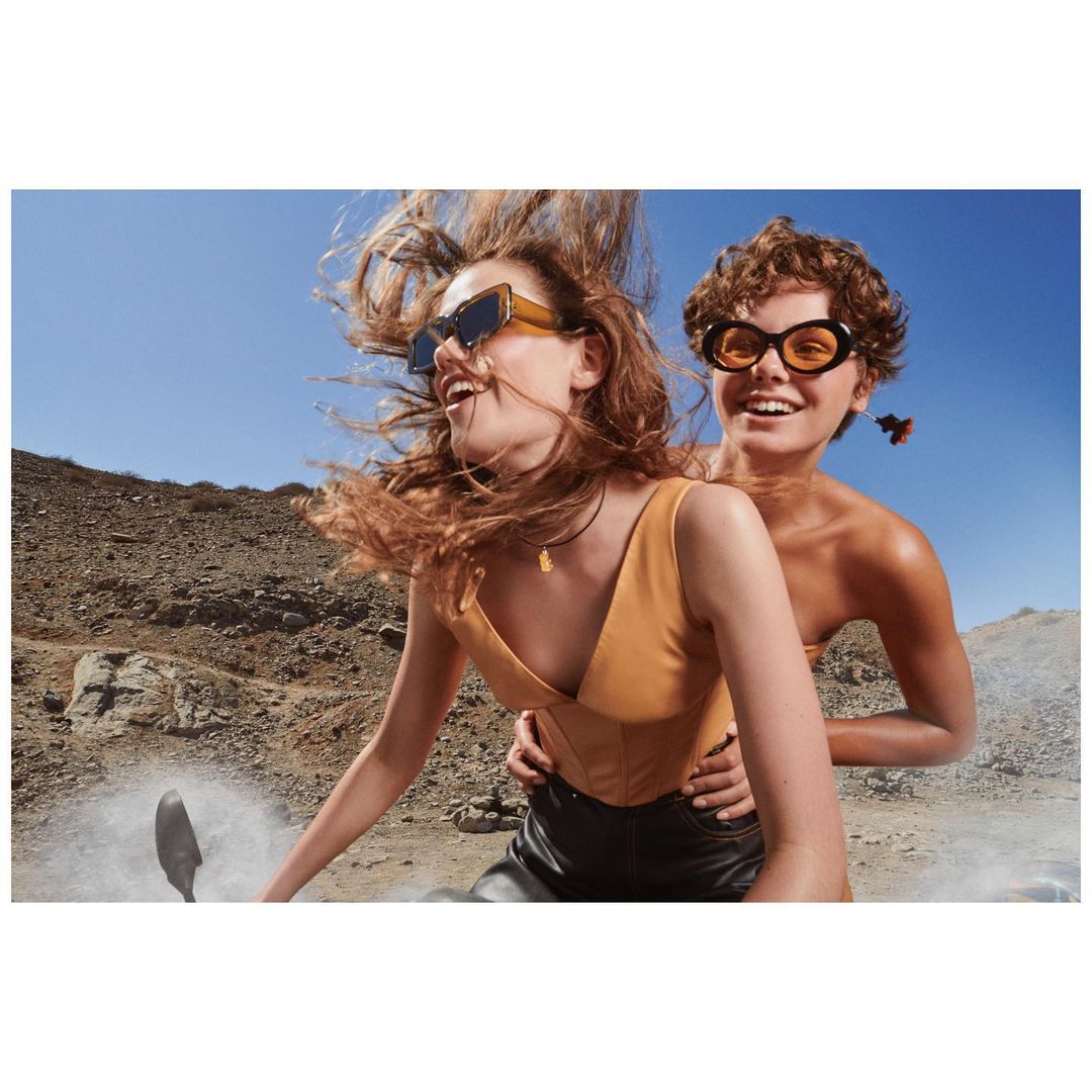 Two women smiling and riding a scooter on a desert road, both wearing sunglasses and fashionable clothing on a sunny day.